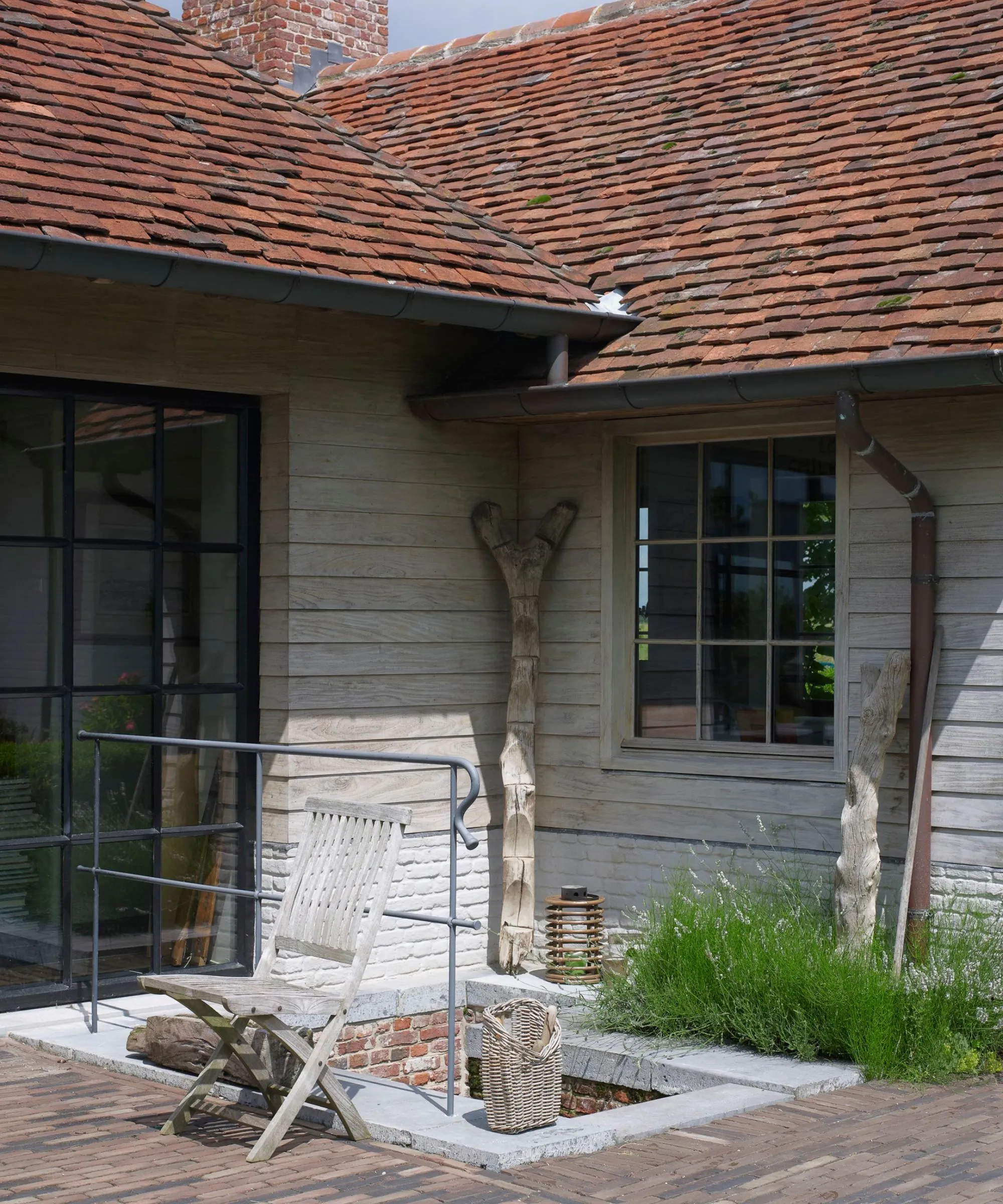 Una casa rivestita in legno con finestre crittali, tegole rosse, una sedia in legno, una grondaia e un tubo di scarico.' loading='lazy' title=