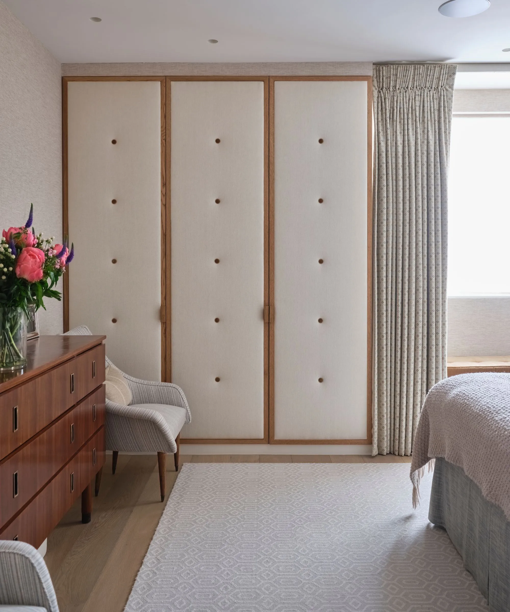 Armário acolchoado em creme e madeira em um quarto com tapete creme, aparador de madeira com vaso de flores rosa brilhante e cadeira cinza.' loading='lazy' title=