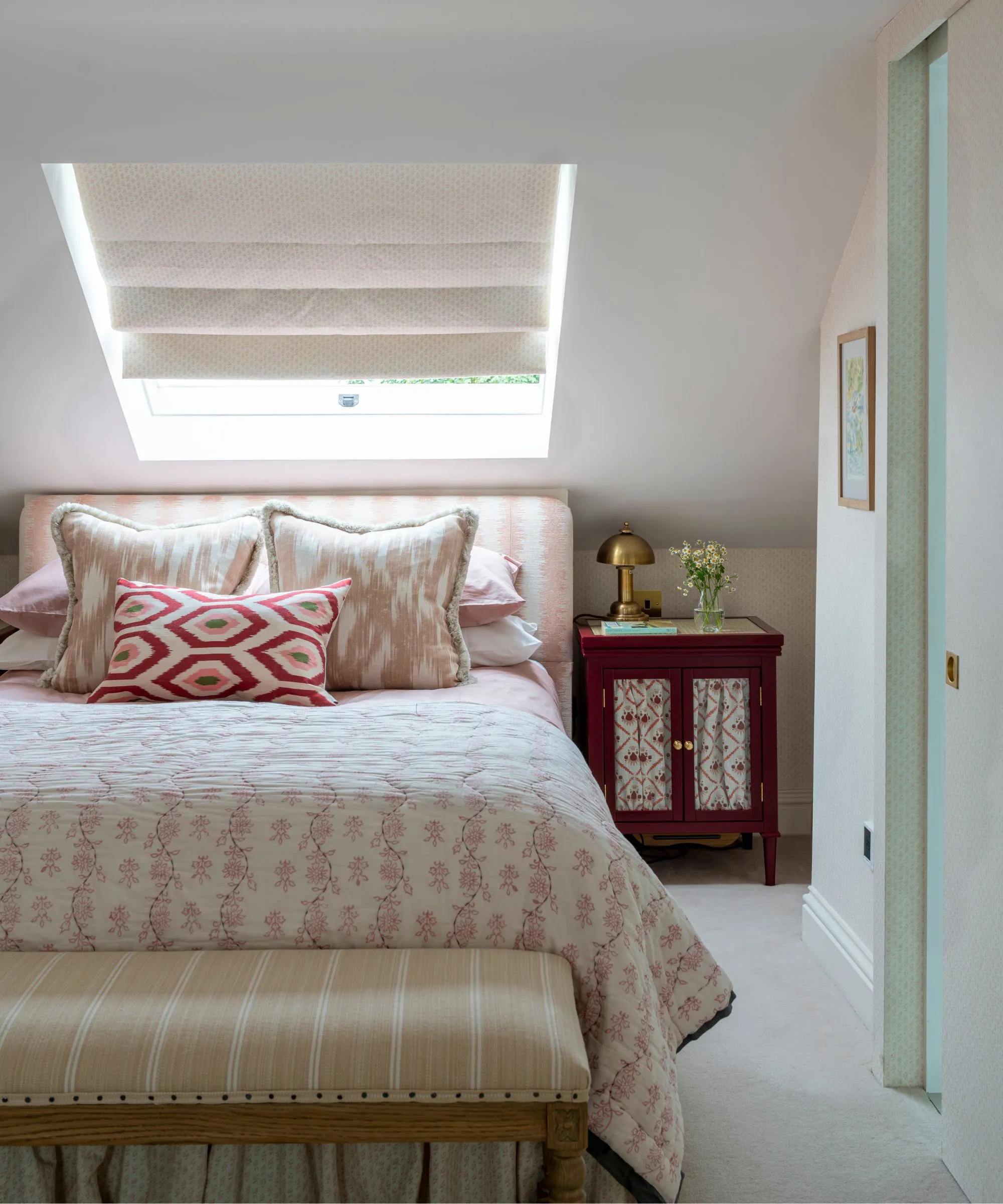 Um quarto branco no sótão com uma grande cama de casal com almofadas estampadas em rosa e branco e cabeceira, uma mesa de cabeceira marrom otomana listrada com abajur dourado e persiana branca em uma janela inclinada acima da cama.' loading='lazy' title=