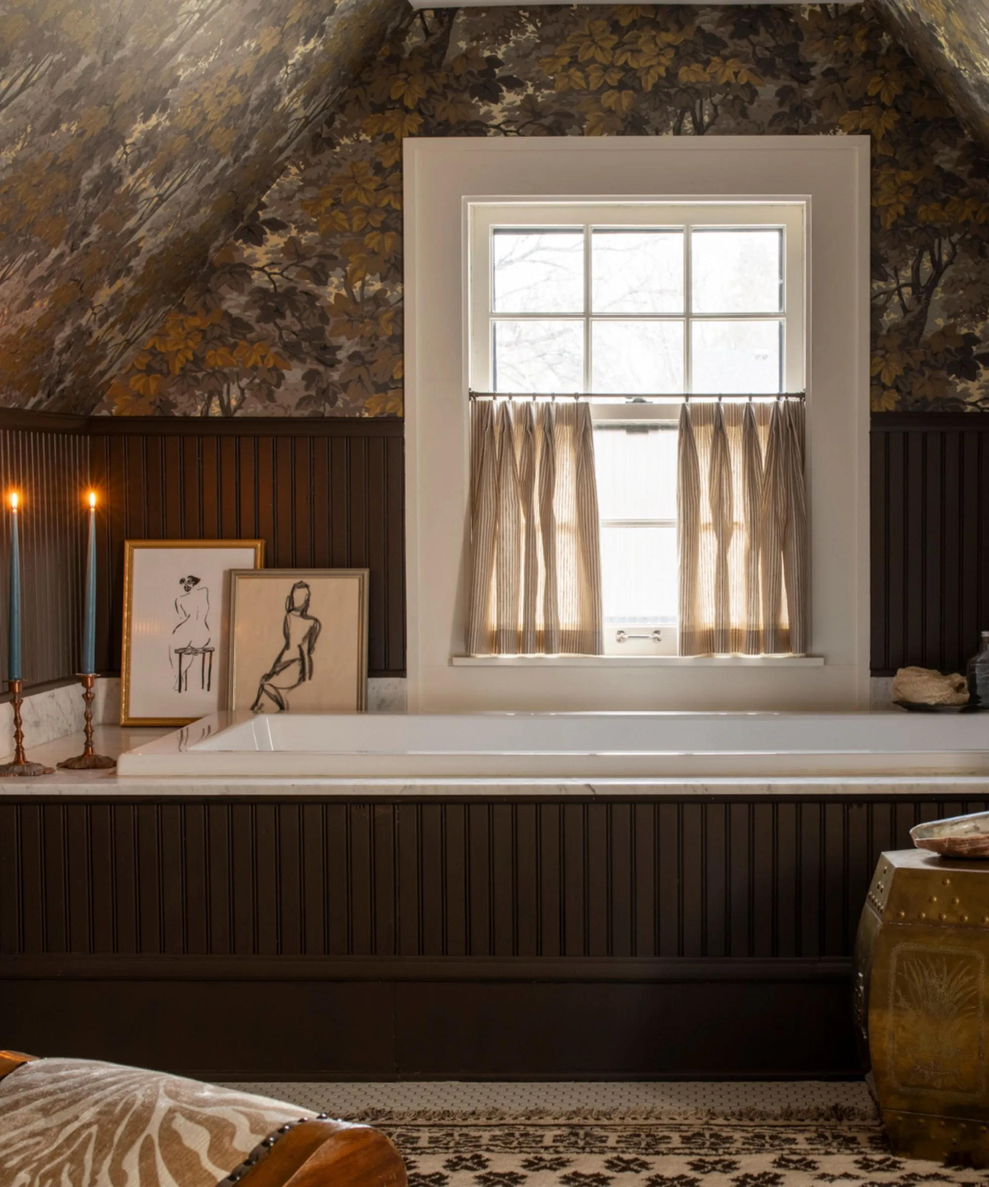 Stimmungsvolles Loft-Gästebad mit dunkelbrauner Holztäfelung und Badewannenverkleidung, kleines Fenster mit Café-Vorhang, botanische Tapete, dekoriert mit einem Teppich, einem Hocker, Kunstwerken, Kerzen und einem kleinen Stuhl' loading='lazy' title=
