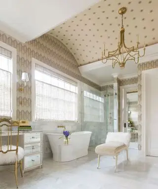 Grande banheiro luxuoso com teto e paredes com papel de parede creme, banheira branca, chuveiro de vidro, assento, piso de mármore e azulejos, lustre dourado,