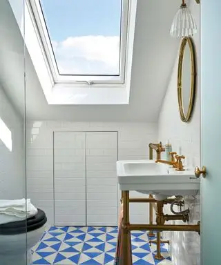 Banheiro branco brilhante com paredes e teto pintados de branco, piso geométrico azul e branco, claraboia no teto inclinado, pia branca tradicional com tubulação de metal exposta, espelho arredondado de metal e aplique de metal e vidro