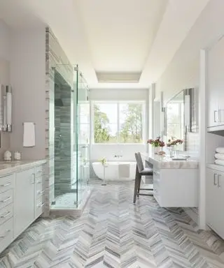 Banheiro amplo com teto embutido branco, piso de cerâmica cinza espinha de peixe com acabamento em mármore, vista para pequena banheira branca, chuveiro com porta de vidro, armários off-white com bancada de mármore, área de estar, paredes de azulejos