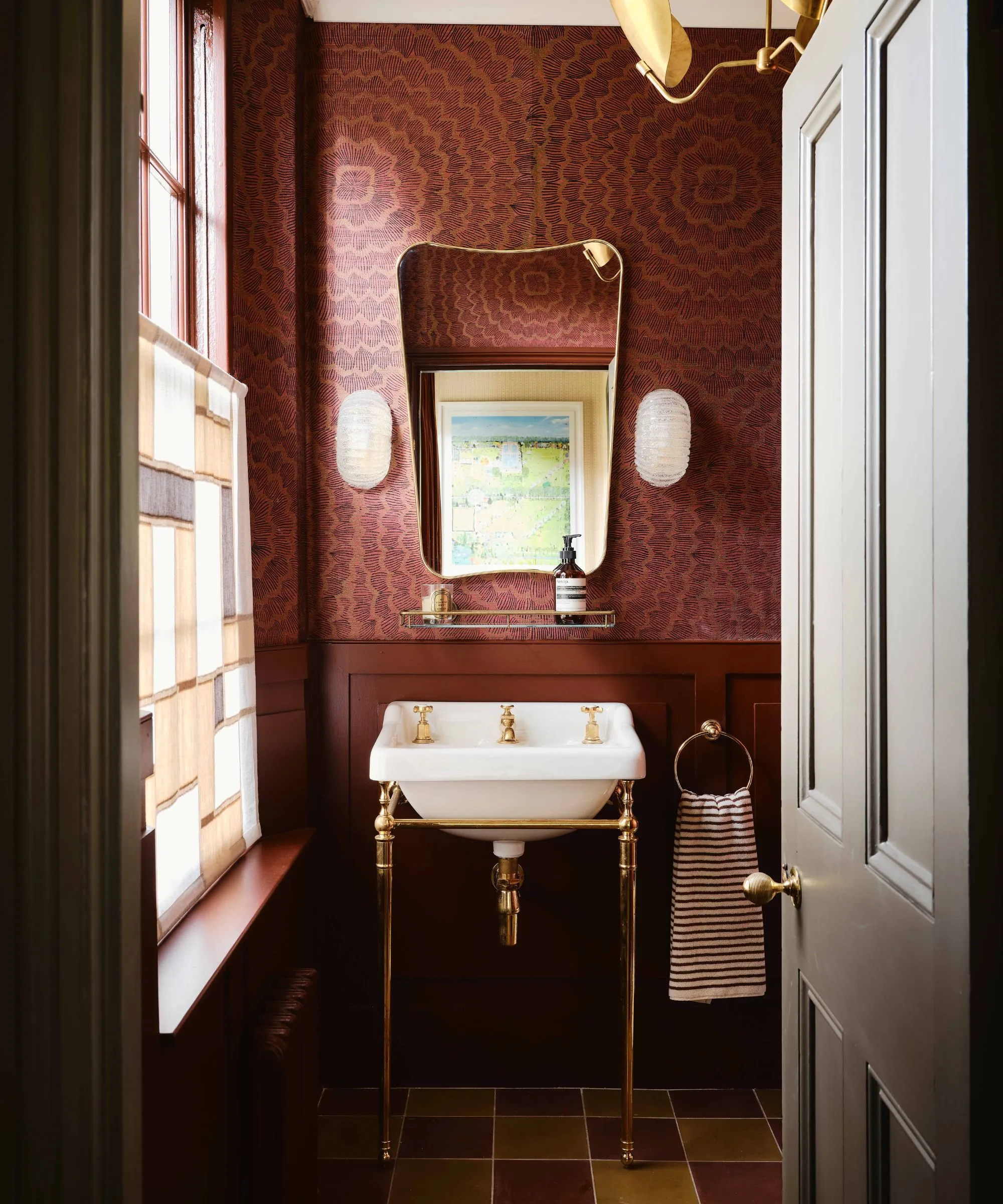 banheiro em pó vermelho escuro com paredes com painéis e papel de parede acima com arandelas de parede vintage espelhadas de meados do século e uma pia tradicional em um suporte de latão' loading='lazy' title=