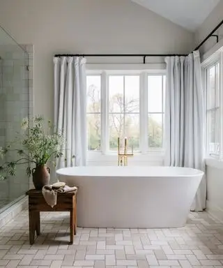 Bagno neutro con vasca bianca, pavimento quadrato coordinato, piastrelle della doccia e tende chiare con ambientazione paesaggistica oltre la finestra