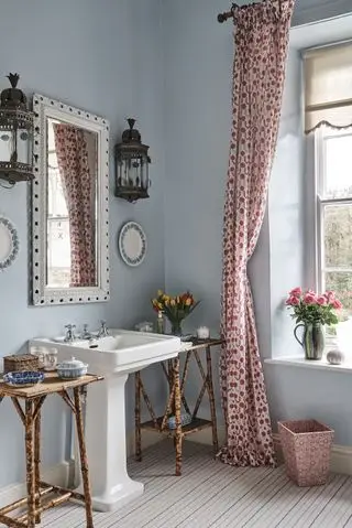 Et eksempel på blå badeværelsesidéer, der viser et pastelblåt badeværelse i tidstypiske omgivelser, gardiner med pink print, et udsmykket spejl, væglamper og sideenheder i rattan
