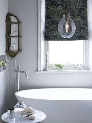 Badezimmer mit freistehender Badewanne mit Pendelleuchte darüber, gemustertem Raffrollo, Beistelltisch und Eckregalen mit weißen Wänden
