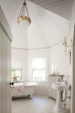 baño blanco con techo abovedado, paredes y techo traslapados, bañera blanca con patas, lavabo con curvas, baldosas hexagonales pequeñas, apliques, radiador, lámpara colgante