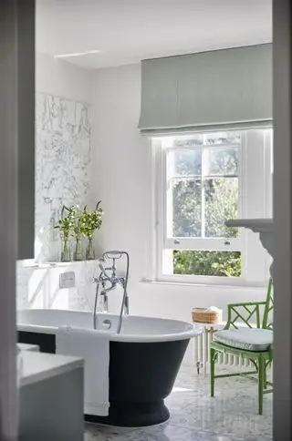 Cuarto de baño blanco con paredes y suelo de mármol, silla verde, persiana verde pálido y encimera enrollable pintada de negro.
