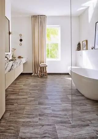 cuarto de baño moderno con bañera blanca, pared de ducha de cristal, tocador de mármol, taburete, cortinas color crema y suelo de vinilo