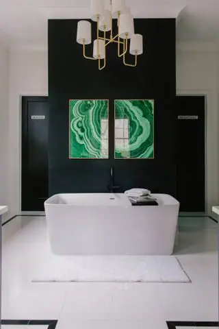 Et rektangulært hvitt badekar på et bad med svarte vegger og hvite gulvfliser, moderne grønt kunstverk på veggen og et gull og hvitt pendellys