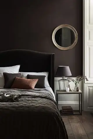 brunt soverom med brun trukket seng, brune vegger, rundt speil, sidebord med glassbasert lampe og brun skjerm. brune og okerfargede tepper, puter