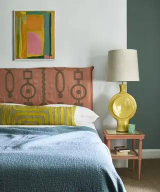 quarto colorido com cabeceira bordada rosa e luz de cabeceira amarela, colcha azul e obras de arte coloridas
