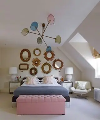 Un ejemplo de ideas de luces de techo para dormitorios que muestran una cama negra frente a espejos decorativos de pared dorados y debajo de una gran lámpara de techo azul, rosa y verde.