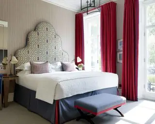 Ein Beispiel für Ideen für Schlafzimmervorhänge, das ein Schlafzimmer mit roten Vorhängen mit blauem Rand und einem großen Bett mit einem auffälligen gepolsterten Kopfteil zeigt