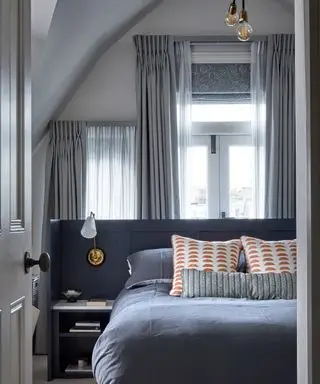 Ein Schlafzimmer mit einem blauen Bett und Fenstern mit zusätzlichen transparenten Vorhängen und einer gemusterten Jalousie