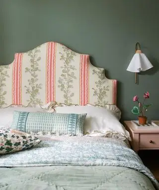 Paredes pintadas de verde no quarto, cabeceira grande estofada com tecido floral, roupa de cama estampada em azul, verde e branco, mesa ao lado de madeira com luminária de parede com abajur de tecido