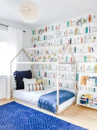 Dormitorio de niños con papel tapiz de estantería, cama con armazón de casa, alfombra azul y suelos de madera.