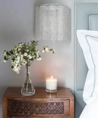 Una pequeña vela sobre una mesita de noche de madera oscura junto a un jarrón de cristal con flores blancas