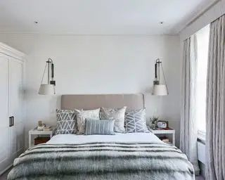 Doppelbett mit grauen und silbernen Kissen und Bettüberwürfen, Einbauschränken auf der linken Seite und Schiebefenster mit langem Vorhang auf der rechten Seite.