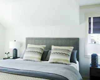 Hellweißes Schlafzimmer mit blauen Akzenten, dunkelgrauem gepolstertem Kopfteil, blau-weißer Bettwäsche, passenden Nachttischlampen und passenden blauen und weißen Jalousien