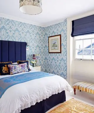 Schlafzimmer in Hellblau und Weiß dekoriert, mit blau-weißer Blumentapete, gepolstertem blauen Kopfteil, weißer Bettwäsche mit blauem Überwurf, mehrfarbigen Zierkissen, blauen Jalousien, gerahmten Kunstwerken an der Wand