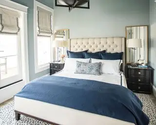Blaues Schlafzimmer mit hellblau gestrichenen Wänden, cremefarbenem gepolstertem Kopfteil, weißem Leinen mit blauem Überwurf und Kissen, Nachttischen aus dunklem Holz, passenden Nachttischlampen mit weißen Schirmen, zwei großen Spiegeln auf beiden Seiten des Bettes, Teppich mit Tiermotiv