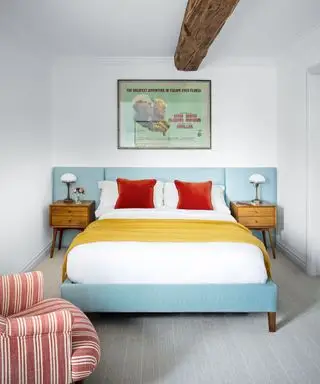 Schlafzimmer mit Sichtbalken, Doppelbett mit türkisfarbenem, übergroßem Kopfteil und rot-weiß gestreiftem Sessel auf Wollteppich.