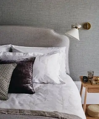 Entspannendes und stilvolles Schlafzimmer mit grauer Strukturtapete, weißer und brauner Bettwäsche. Ein Doppelbett und eine schräge Wandleuchte.