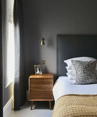 Eine graue Schlafzimmeridee mit dunklen Wänden, einem Beistelltisch aus warmem Holz und einem ockerfarbenen Überwurf auf dem Bett