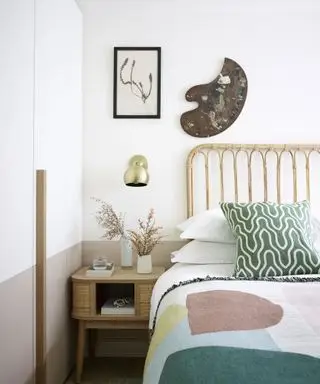 Quarto branco claro com metade inferior da parede pintada em marrom claro, cama estilo bambu, roupa de cama branca com almofadas estampadas verdes, manta em tons pastéis com formas geométricas, mesa de cabeceira de madeira com pequenos vasos de flores, aplique metálico acima, obras de arte emolduradas e paleta de pintores montada acima da cama,