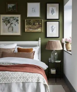 Quarto pintado de verde com parede de galeria acima da cama, moldura branca com linho branco, almofadas e mantas em paleta de cores terrosas, mesinha de cabeceira de metal com abajur com base de vidro, abajur de tecido, piso de pedra, tapete creme