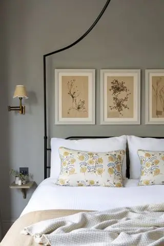 dormitorio gris con cama de hierro negro, obras de arte, cojines florales, manta de avena, pequeña mesita de noche montada en la pared y lámpara de pared