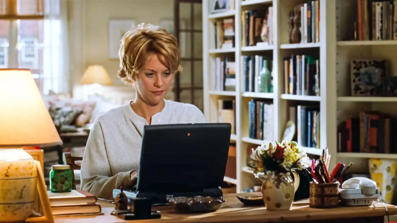 Meg Ryan em You Get Mail (1998) Filme ficou em uma mesa com um laptop com estantes de livros atrás dela e uma lâmpada de mesa' loading='lazy' title=