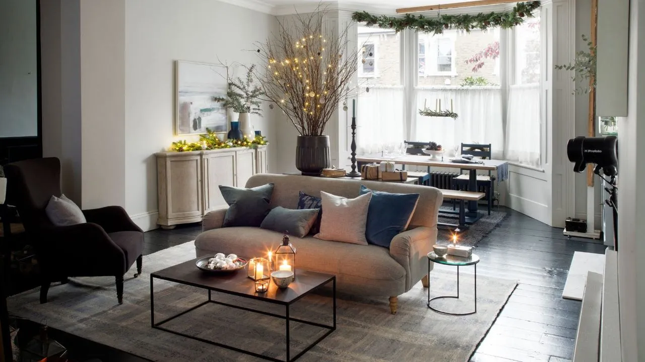 아늑한 축제 분위기의 거실에는 커다란 크림색 소파, 커피 테이블, 불이 켜진 양탄자, 갈색 덮개를 씌운 의자, 흰색 커튼이 달린 대형 돌출형 창문 아래 식탁이 있습니다. 방 중앙에는 불이 붙은 마른 나뭇가지와 키가 큰 촛대가 있는 커다란 꽃병이 있습니다. 창문 위에는 축제용 녹지가 걸려 있고, 오른쪽에는 꼬마전구와 축제 잔가지가 담긴 꽃병, 예술작품이 있는 흰색 사이드 테이블이 있습니다.' loading='lazy' title=