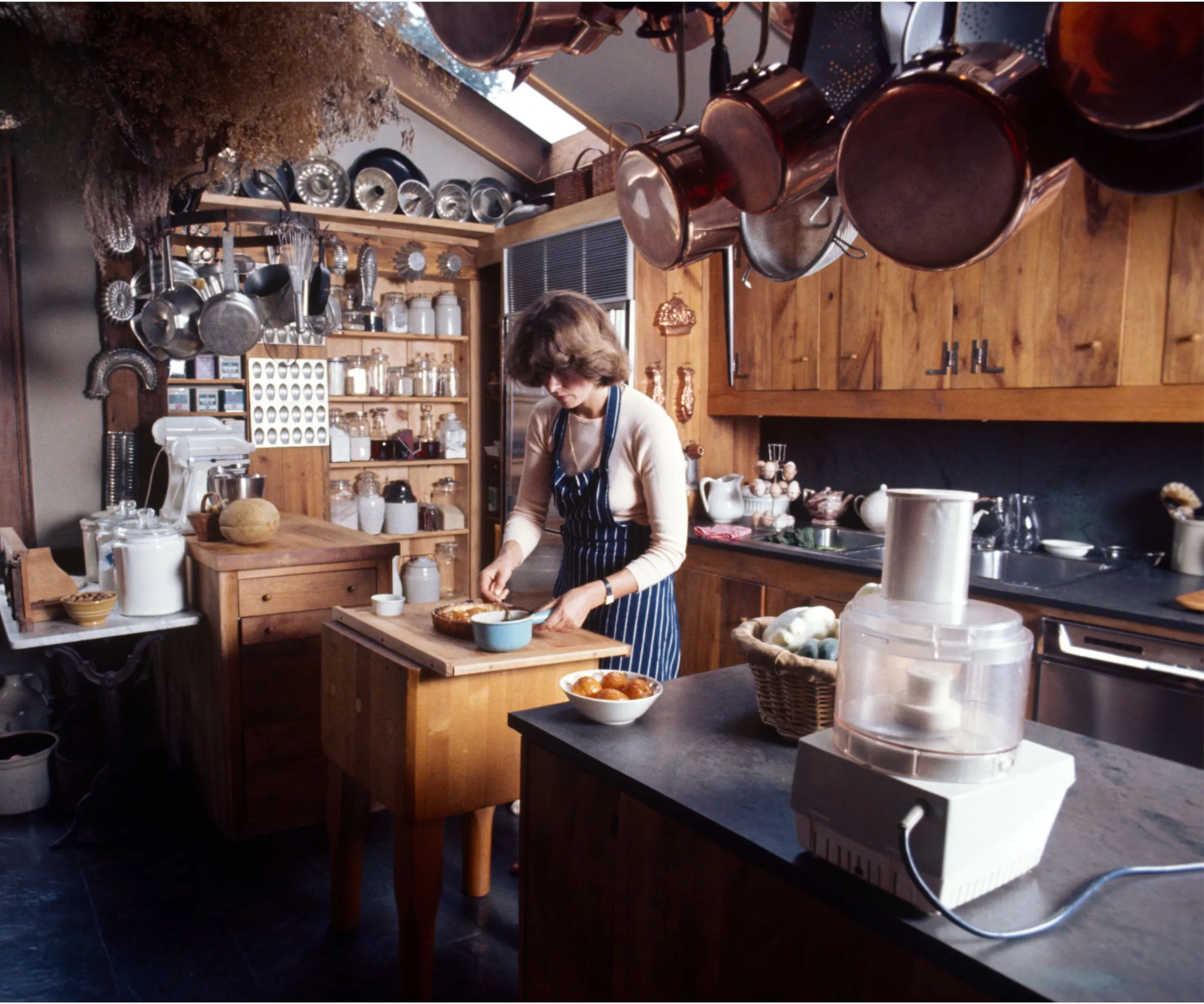 Martha Stewart w swojej wiejskiej kuchni z drewnianymi szafkami kuchennymi, miedzianymi patelniami wiszącymi na poręczowej spiżarni ze słoikami i miedzianymi foremkami' loading='lazy' title=