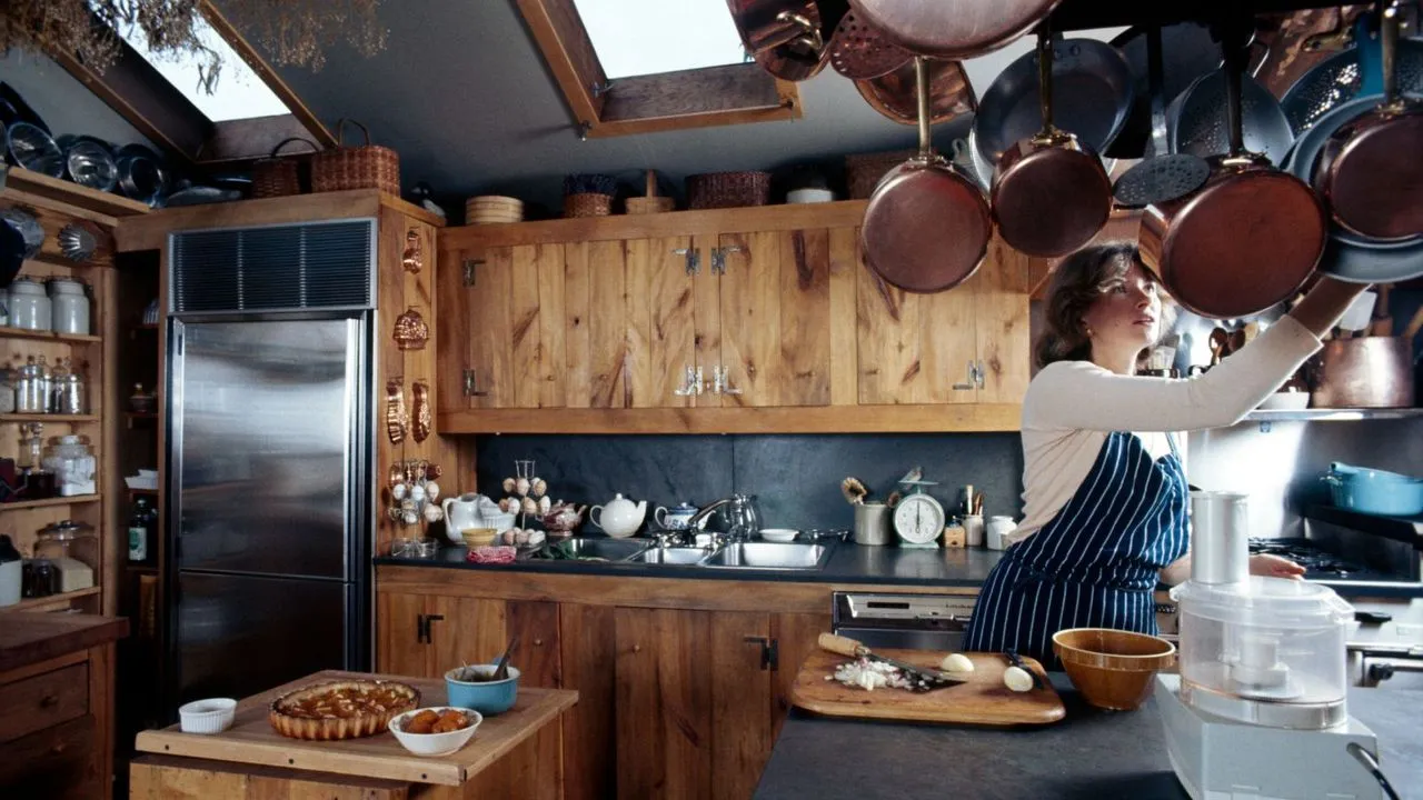 Martha Stewart vo svojej kuchyni, ktorá obsahuje drevené skrinky, medený riad a otvorené police plné pohárov a medených nožov' loading='lazy' title=