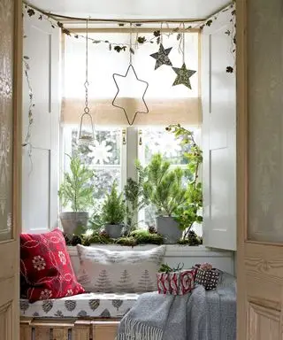 Ideen für weihnachtliche Fensterdekoration mit Sterndekorationen, die über einem Fensterplatz mit roten Kissen und skandinavischen Decken hängen