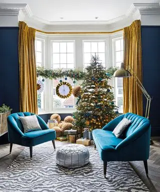 Ideen für weihnachtliche Fensterdekoration in einem blauen Wohnzimmer mit gelben und blauen Dekorationen auf einer Laubgirlande, passend zur Weihnachtsbaumdekoration