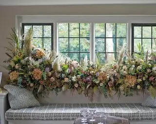 Weihnachtsdeko-Ideen für Fenster mit einem großen Blumenarrangement in Pastellfarben, das sich über die gesamte Länge eines Fensterbretts über einer Bank erstreckt