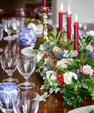Mesa de comedor navideña con follaje, velas rojas y cristalería.