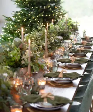 Gran mesa de comedor navideña decorada con follaje verde, velas, vajilla y árbol de Navidad al fondo