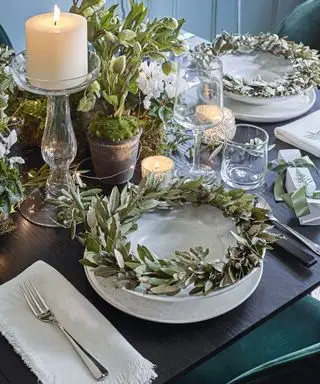 Un lugar para Navidad con velas blancas, candelabros de cristal, coronas de hojas verdes plateadas y macetas de vegetación, hiedra y musgo.