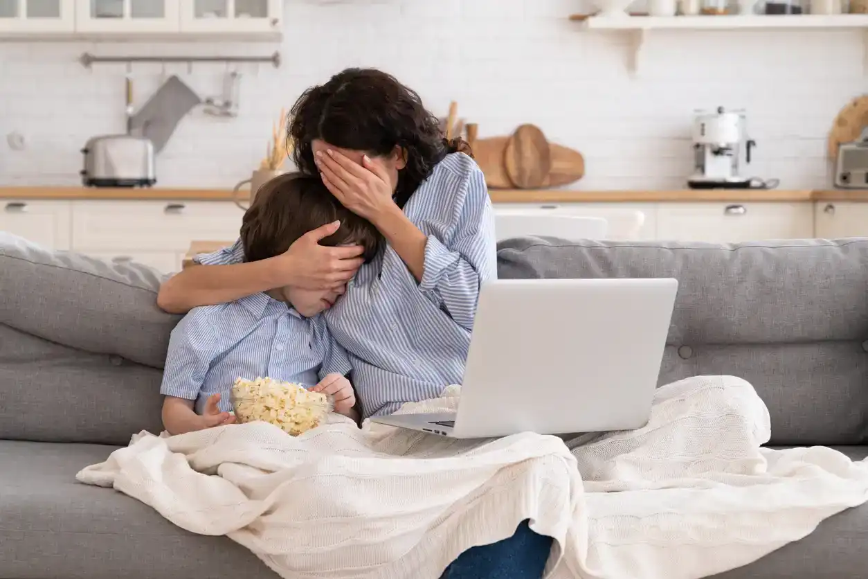 Frightened mom and little kid son with bowl of popcorn watching scary scene in movie closing their eyes, sitting on sofa at home. Mother hugs the child, watches a horror movie, feels fear.