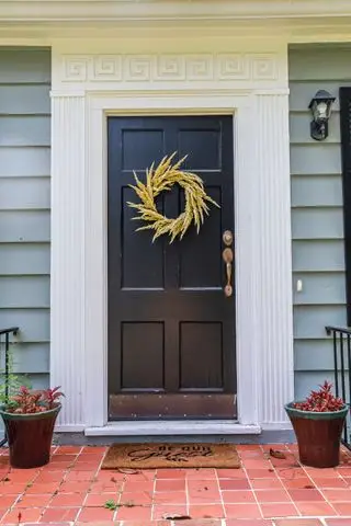 una puerta delantera negra y una corona en una pequeña casa estilo cabaña azul recientemente renovada con escalones de ladrillo