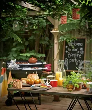 Udendørs spisningsideer med et Bonfire Night-festskilt på en tavle, grill og klistrede æbler og drinks på et bord, med lanterner hængende fra løvtag