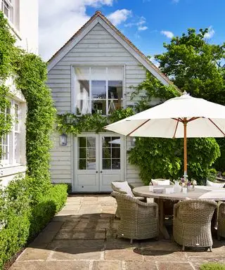 Et rundt udendørs spisebord med rattanstole og en hvid parasol foran et hvidt hus med løv