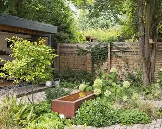 Kleiner Garten mit Ziegelmauer, Ziegelboden, Wasserbehälter aus Metall, Blumen, Büschen, Bäumen