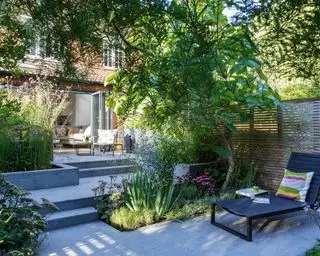 Eine schattige, lange Gartenidee mit grauem Pflaster und Stufen sowie einer schwarzen Sonnenliege.