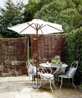 Ein Beispiel für lange Gartenideen mit Korbschirmen und Hecken, die einen Terrassenbereich mit weißem Metalltisch und Stühlen unterteilen.